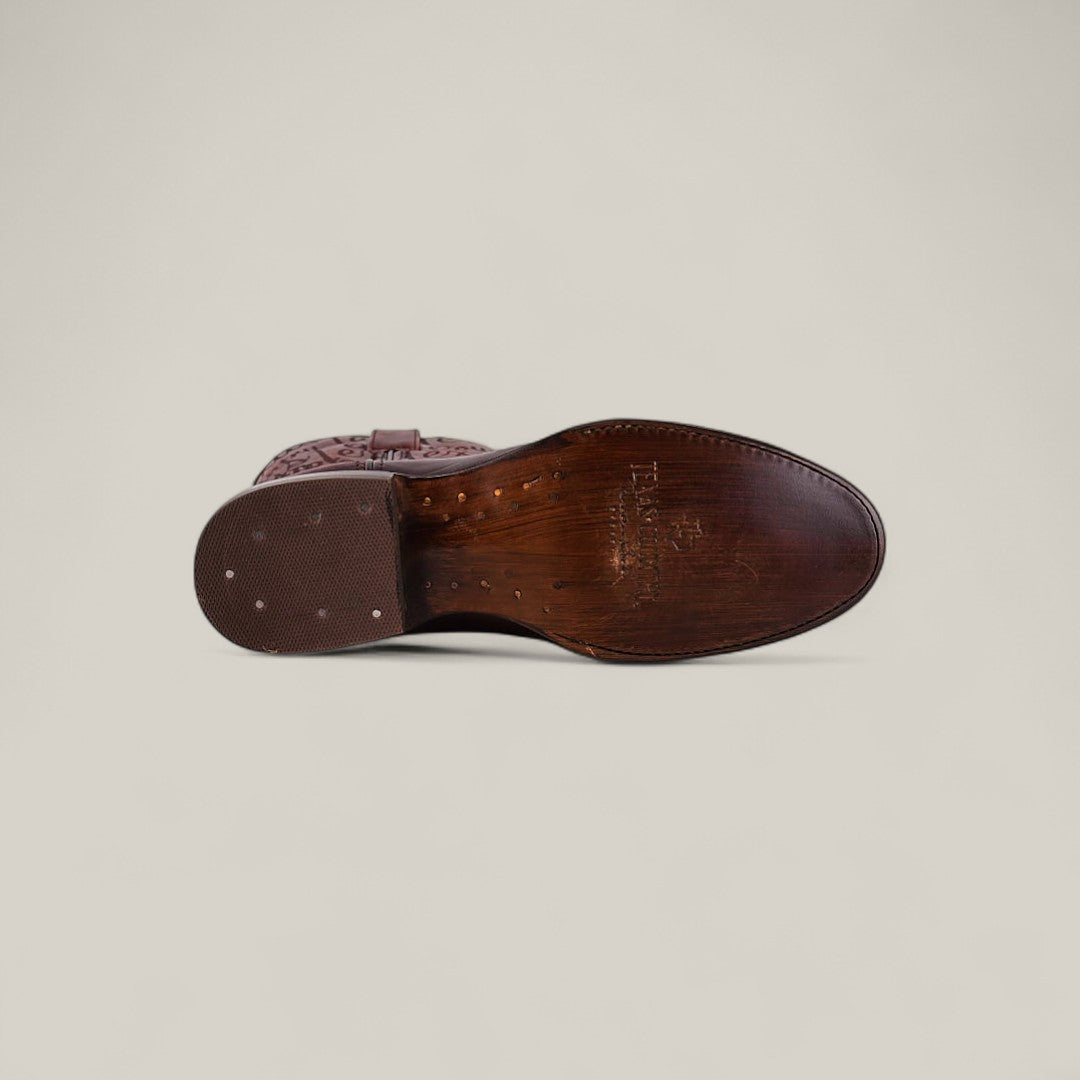 The Wyatt - Cogñac - Roper Toe is a brown leather dress shoe, shown from the bottom with a smooth, polished sole, small metal nails at the heel, and embossed branding near the arch. Set against a light beige background.