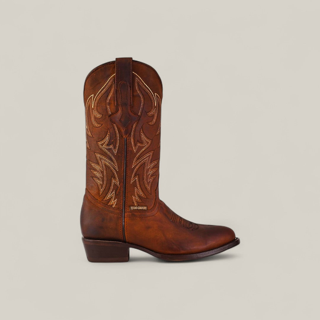 The Dutton - Brick - Roper Toe boot, crafted from brown cowhide with decorative stitching, a low heel, and a pointed toe, is displayed against a light background, highlighting its classic leather boot style.