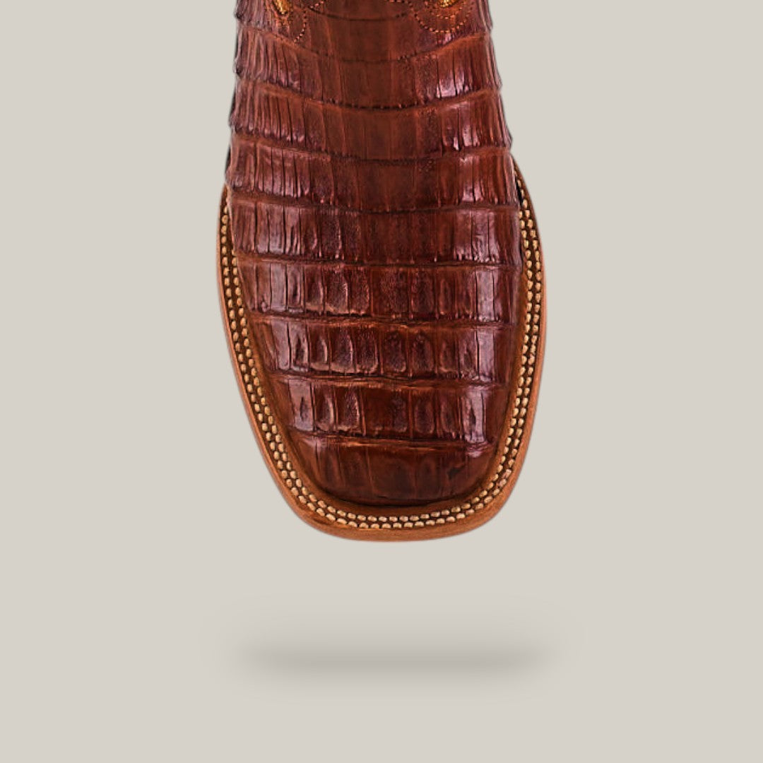 A close-up of the Exotic Caiman Belly Tail - Brandy - Square Toe showcases its caiman leather with a crocodile skin texture, detailed edge stitching, and brandy brown color, set against a light beige background.