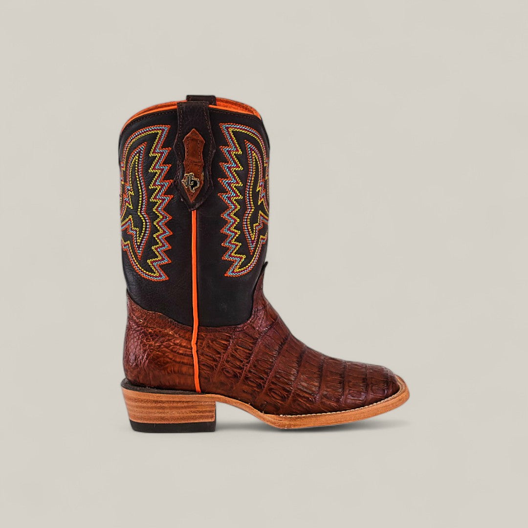 The Exotic Caiman Belly Tail Cognac Square Toe Kids Boot features a brown textured leather foot, black shaft with colorful zigzag stitching, orange piping, and a wooden heel, displayed on a plain light beige background.