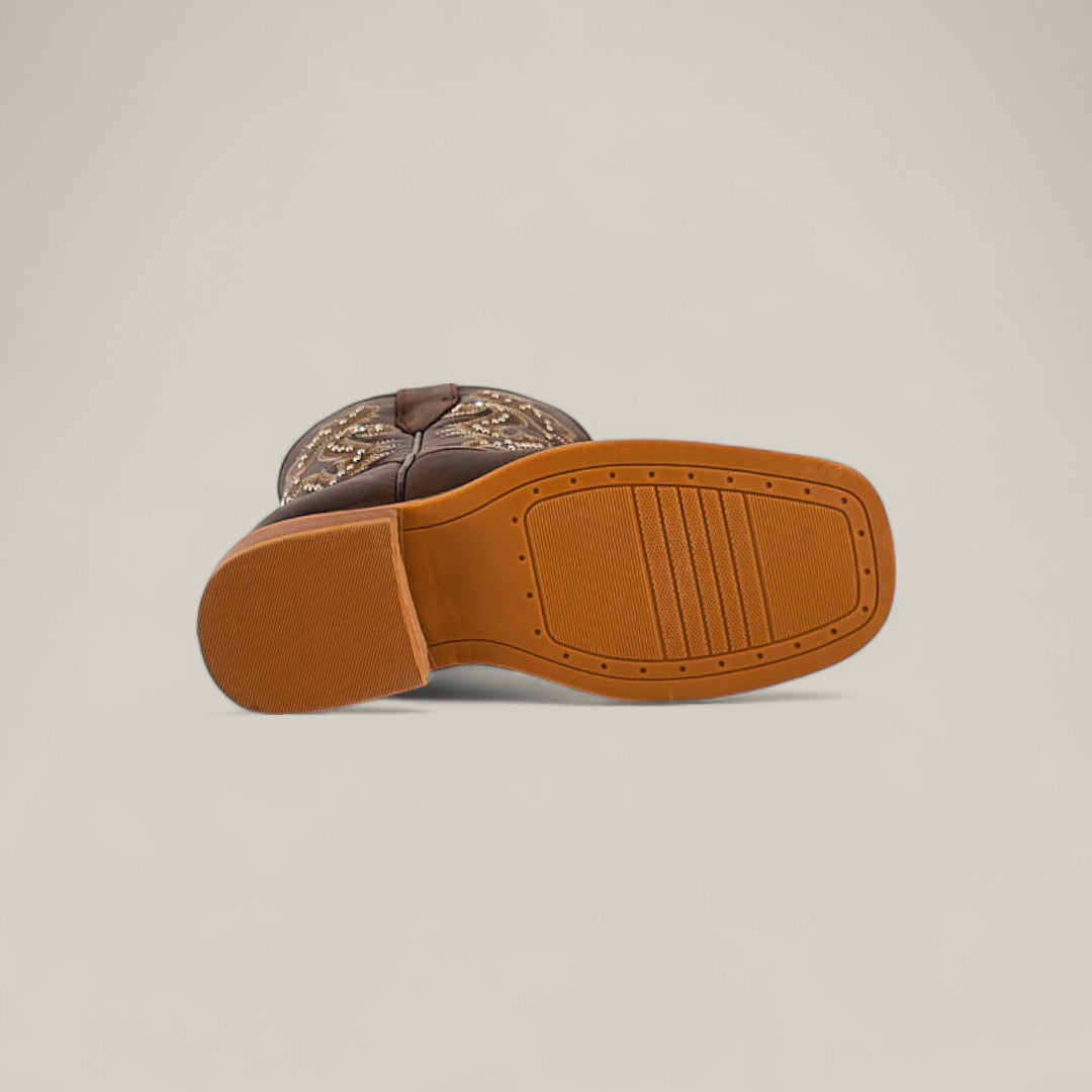 The Britany Brown - Square Toe - Kids Boots is shown on its side, revealing a tan rubber sole with textured grip and horizontal grooves. The brown upper has lighter decorative stitching, emphasizing its quality craftsmanship.