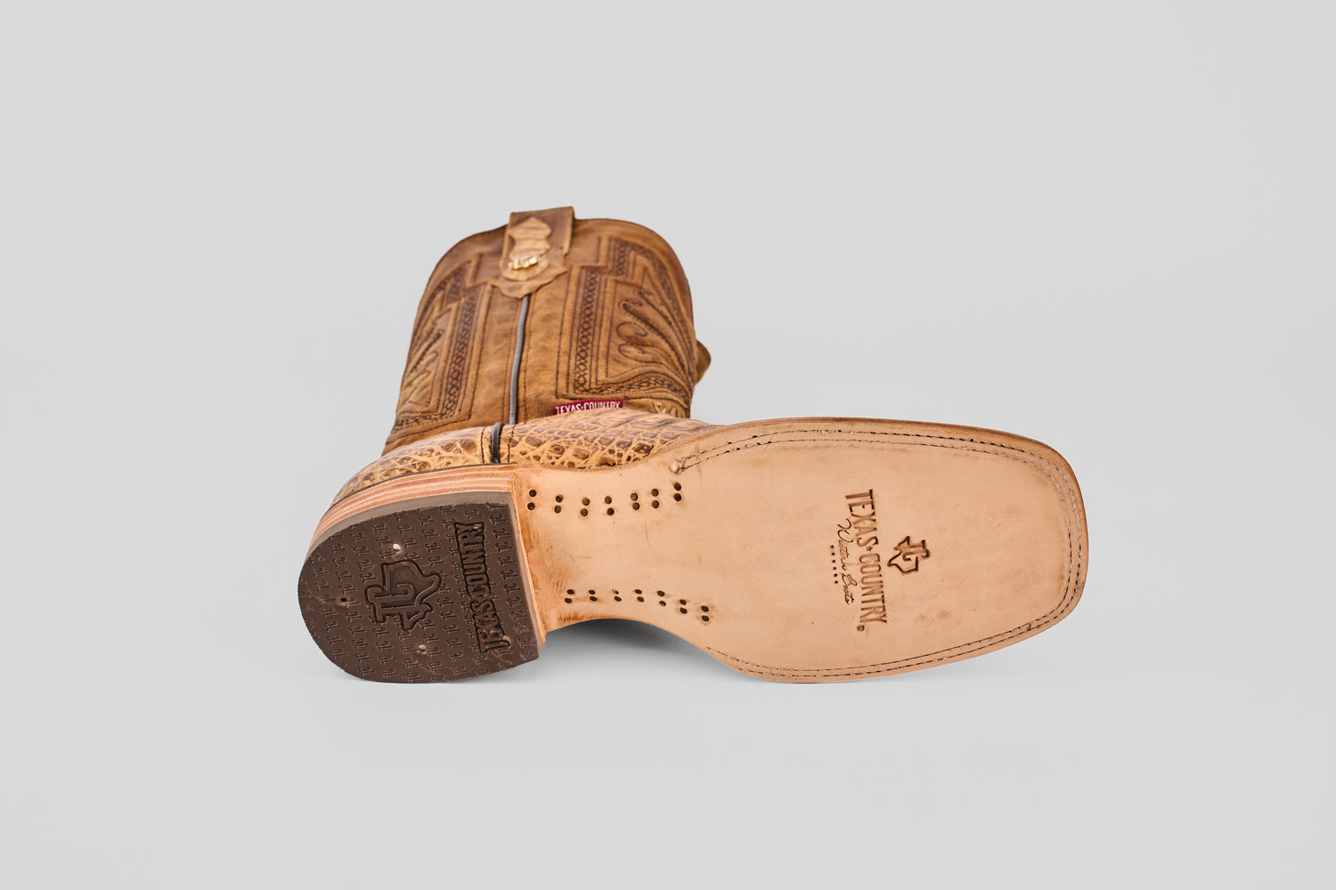 The Exotic Caiman Belly - Antique - Square Toe boot is shown on its side, highlighting the detailed shaft embroidery and stamped TEXAS COUNTRY logo on the tan sole. The square toe design features a black rubber heel grip with a star motif.