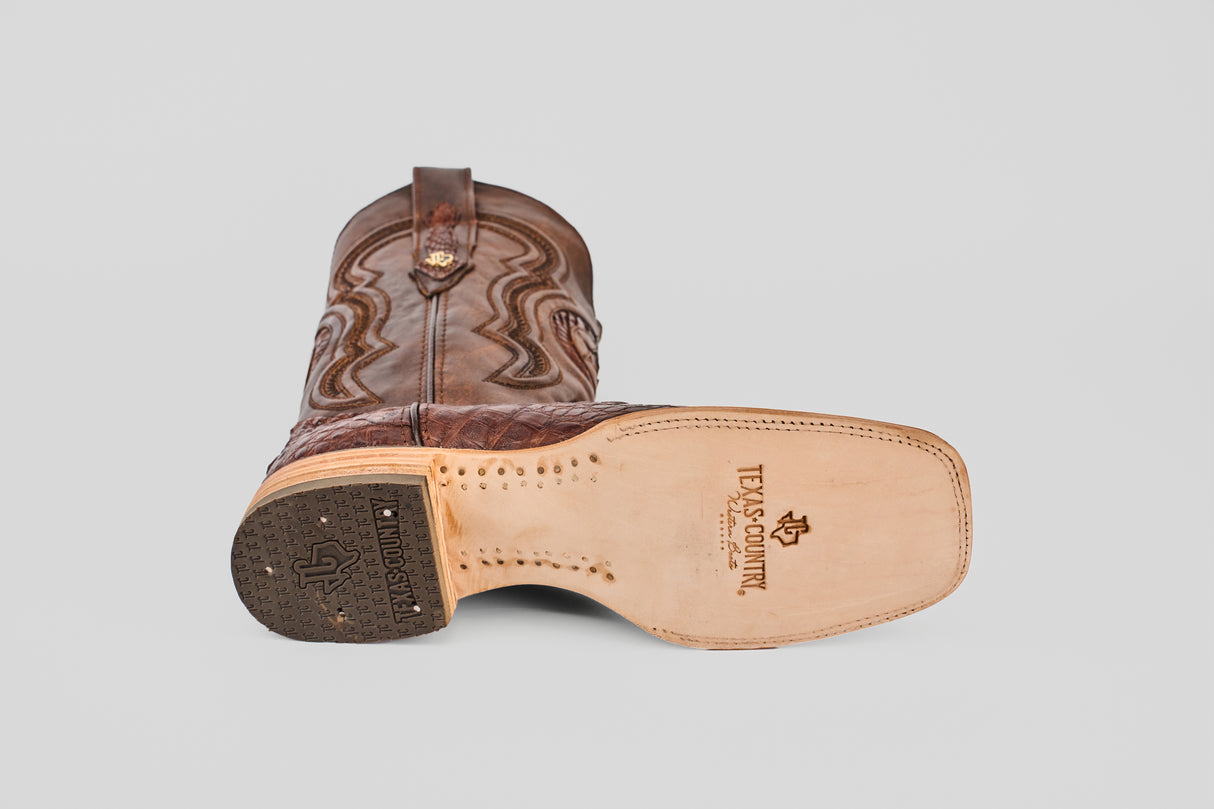 A brown Exotic Caiman Horn Back - Square Toe boot lies on its side, showing the tan sole with visible stitching, tread on the heel, and the Texas Country logo—an iconic style for fans of square toe boots.