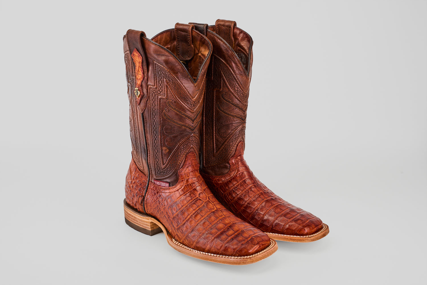A pair of Exotic Caiman Belly - Brandy - Square Toe boots with detailed stitching and a textured alligator pattern are displayed side by side on a light gray background.