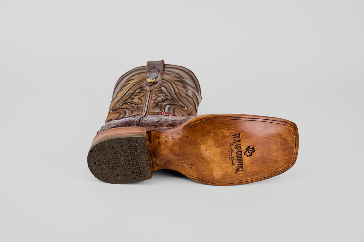 A brown Exotic Caiman Hornback - Brown - Rodeo Toe boot rests on its side, displaying the Texas Country logo stamped on genuine caiman leather, highlighting its premium quality against a plain gray background.