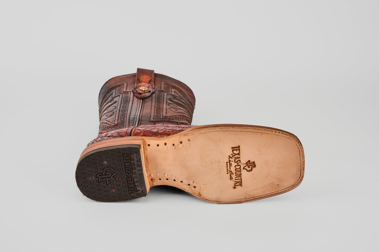 A brown Exotic Caiman Horn Back - Smoke Cogñac Square Toe boot lies on its side, showcasing the Texas Country logo stamped on the leather sole and a decorative upper pattern.
