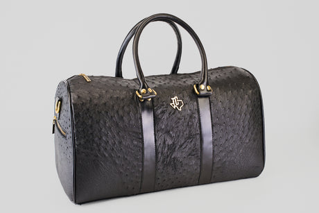 The Exotic Full Quilt Ostrich - Black Travel Bag features gold hardware, a subtle ostrich-textured pattern, and a gold logo on the front, all set against a plain light gray background—your perfect luxurious travel companion.