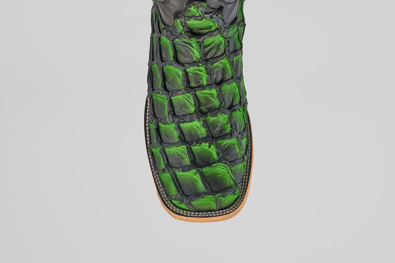 A close-up, top view of the Conor - Swamp Green - Square Toe boot showcases green and black scale-textured leather with visible stitching on a tan sole, all set against a plain white background.