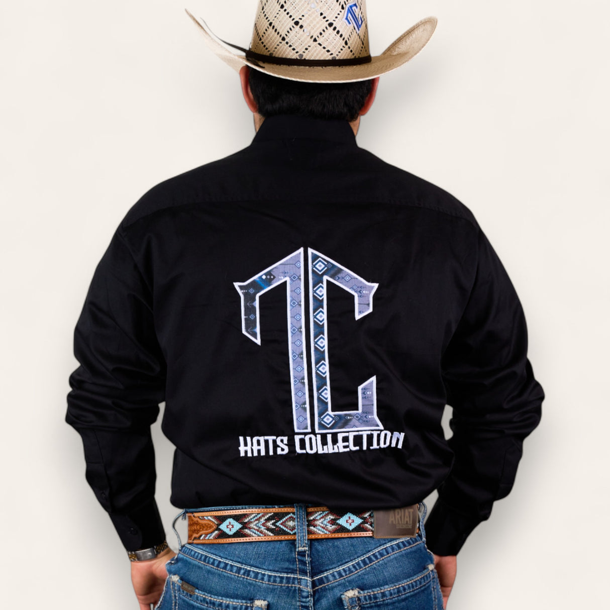 A man in a cowboy hat, black shirt with a large TC Hats Collection logo on the back, and blue jeans wears the TC Hats - Black Blue Aztec belt, featuring a stylish white and blue Aztec design, standing with his back to the camera.