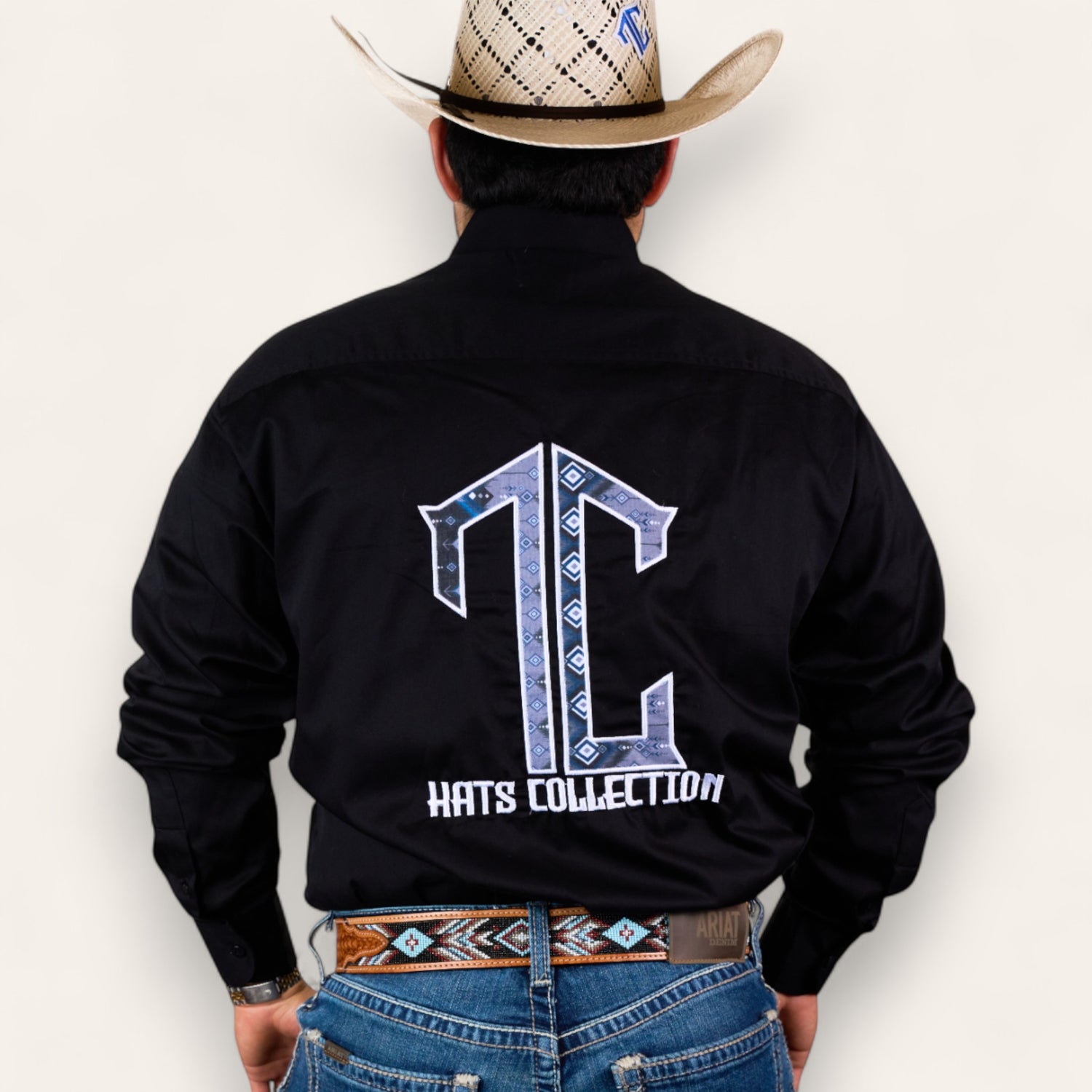 A man in a cowboy hat, black shirt with a large TC Hats Collection logo on the back, and blue jeans wears the TC Hats - Black Blue Aztec belt, featuring a stylish white and blue Aztec design, standing with his back to the camera.