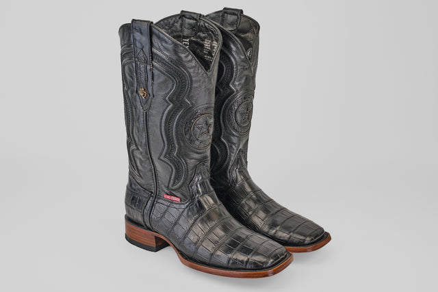 A pair of Exotic Alligator Tail - Black - Square Toe boots, featuring embossed leather, wooden soles, and detailed shaft stitching, displayed on a plain light gray background.