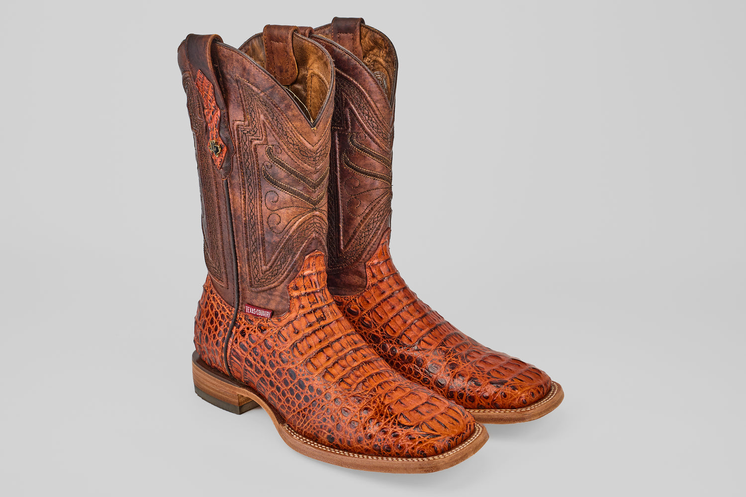 A pair of Exotic Caiman Head - Cogñac - Square Toe boots with patterned, textured caiman leather and decorative stitching on the tall shafts, displayed against a plain light gray background.