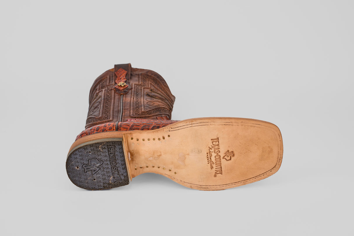 A single Exotic Caiman Head - Cogñac - Square Toe boot lies on its side, displaying the tan leather sole with the Tecovas logo, black heel, caiman texture, and intricate stitching on the upper.