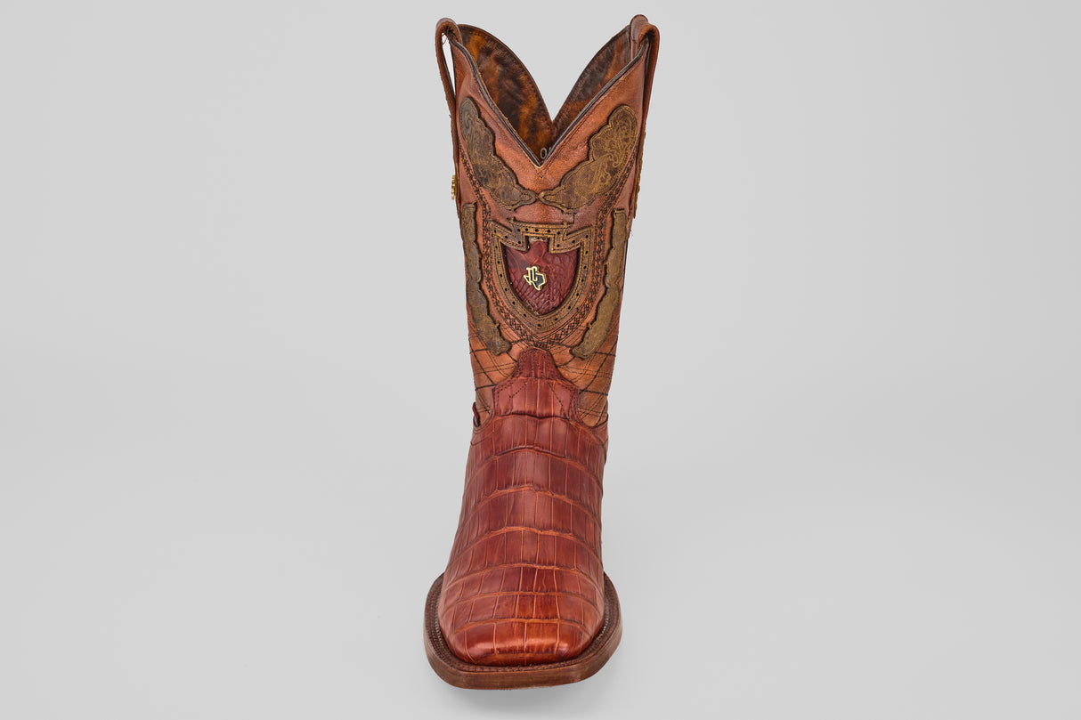 The Exotic Alligator Tail - Brick - Square Toe boot features brown textured leather, decorative stitching, and a red and gold crest on the front shaft, displayed against a plain light background.
