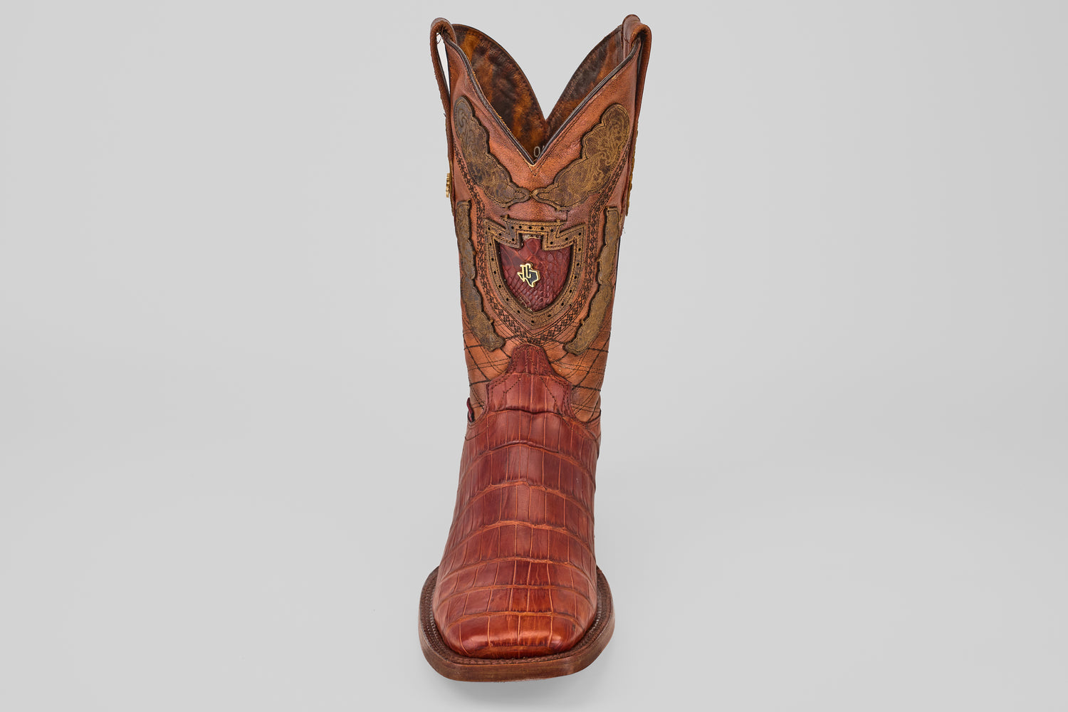 The Exotic Alligator Tail - Brick - Square Toe boot features brown textured leather, decorative stitching, and a red and gold crest on the front shaft, displayed against a plain light background.
