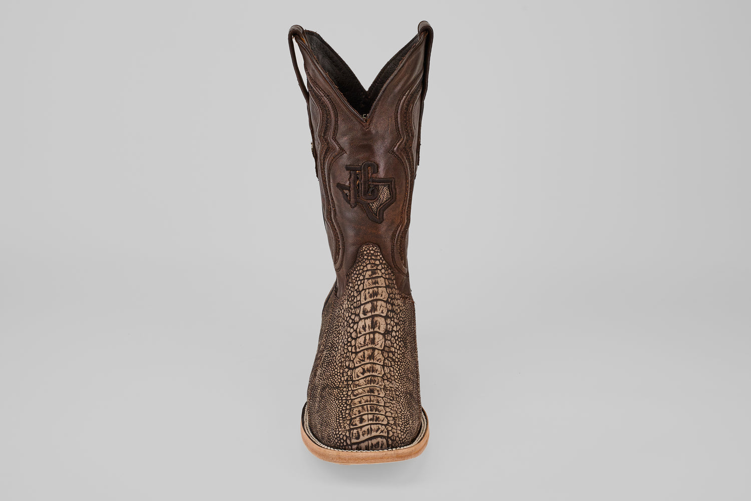 A single Exotic Full Quilt Ostrich Leg - Matte Orix - Square Toe boot, featuring a textured brown patterned front, light tan sole, and decorative shaft stitching, displayed upright on a plain gray background.