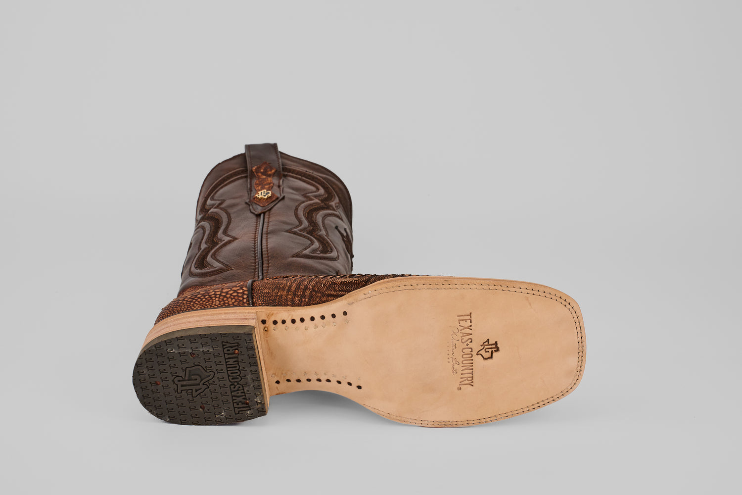 A pair of Exotic Full Quilt Ostrich Leg - Rustic Brick - Square Toe boots, with one boot upright and the other on its side, reveals a smooth tan leather sole stamped with the Texas Country logo and a black textured rubber heel.