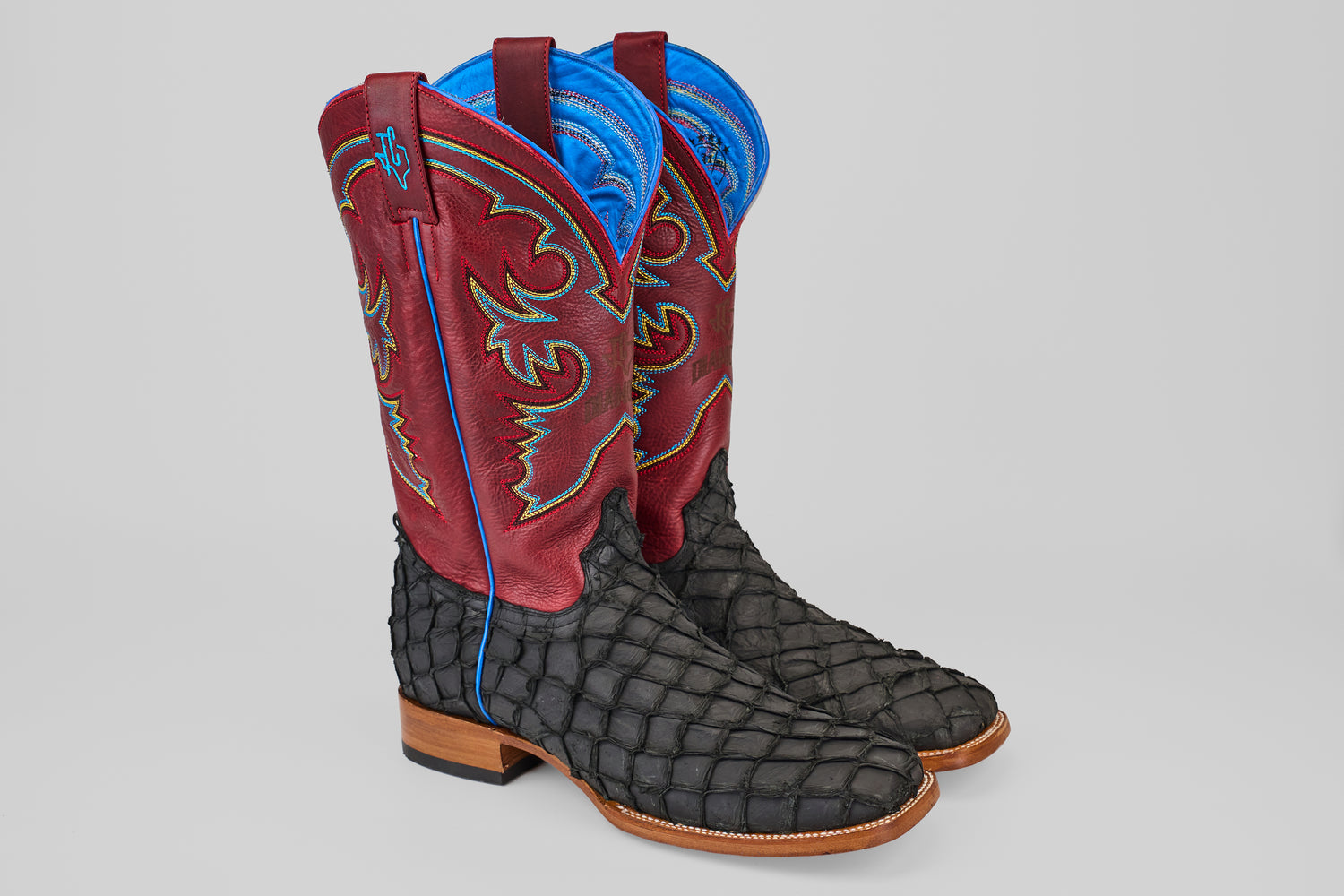 The Dodge - Matte Black - Square Toe boots feature red and blue decorative uppers, textured black Pirarucu leather feet, wooden heels, and light blue stitching accents, displayed against a plain gray background.