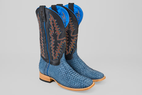 The Kingsley - Blue Jeans - Square Toe boots feature tall, blue snakeskin with black shafts, colorful zigzag stitching, and light brown wooden heels, displayed on a light gray background.