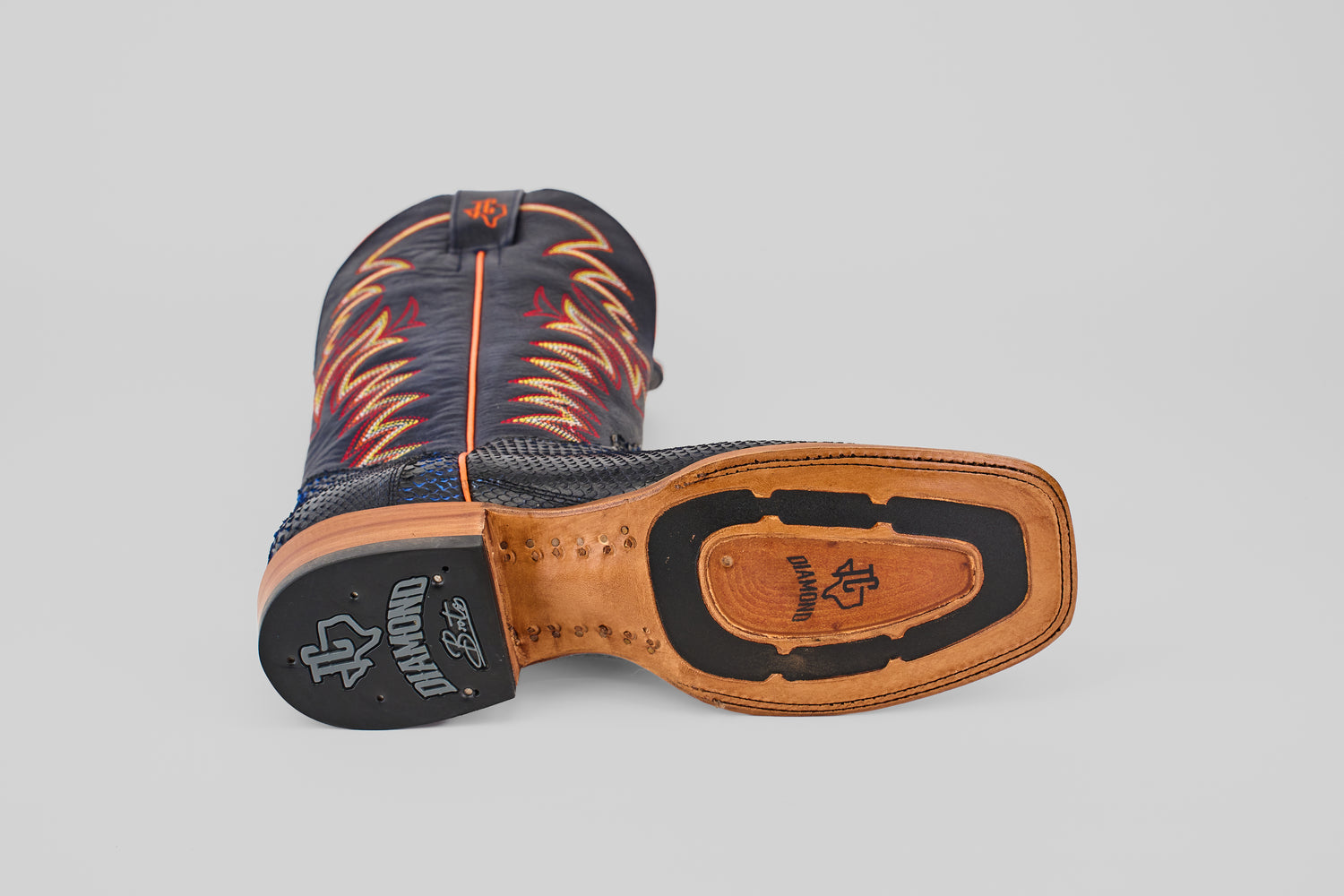 A McGraw - Navy - Square Toe boot from the Diamond Collection lies on its side, displaying a tan leather sole with black rubber heel and logo, plus blue uppers accented by red and yellow flame-like stitching.