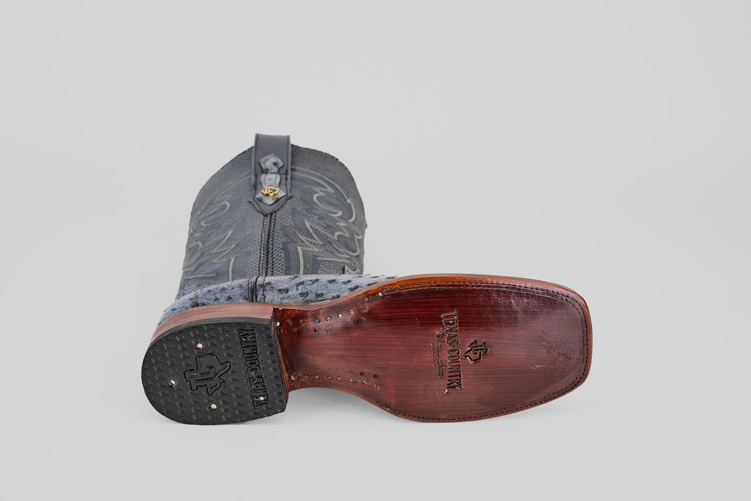 The Exotic Full Quilt Ostrich - Safari Gray - Square Toe boot is displayed on its side, highlighting the decorative stitching on the leather upper and the engraved branding on the wooden sole.