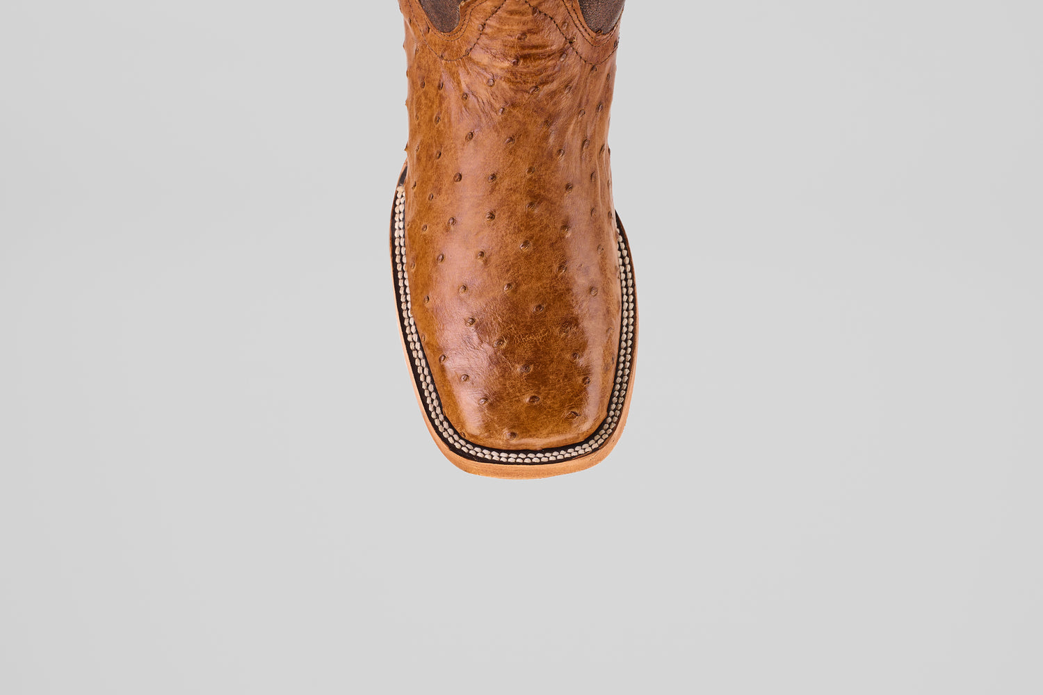 A top view of the Exotic Full Quilt Ostrich - Brandy Brucciato boot features brown leather, a square toe, and decorative edge stitching in classic cowboy style, set against a plain light gray background.