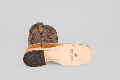 The Exotic Full Quilt Ostrich - Brandy Brucciato - Square Toe boot lies on its side, revealing a beige sole with TEXAS COUNTRY Classic and logo, plus the heel and detailed stitching along the shaft.