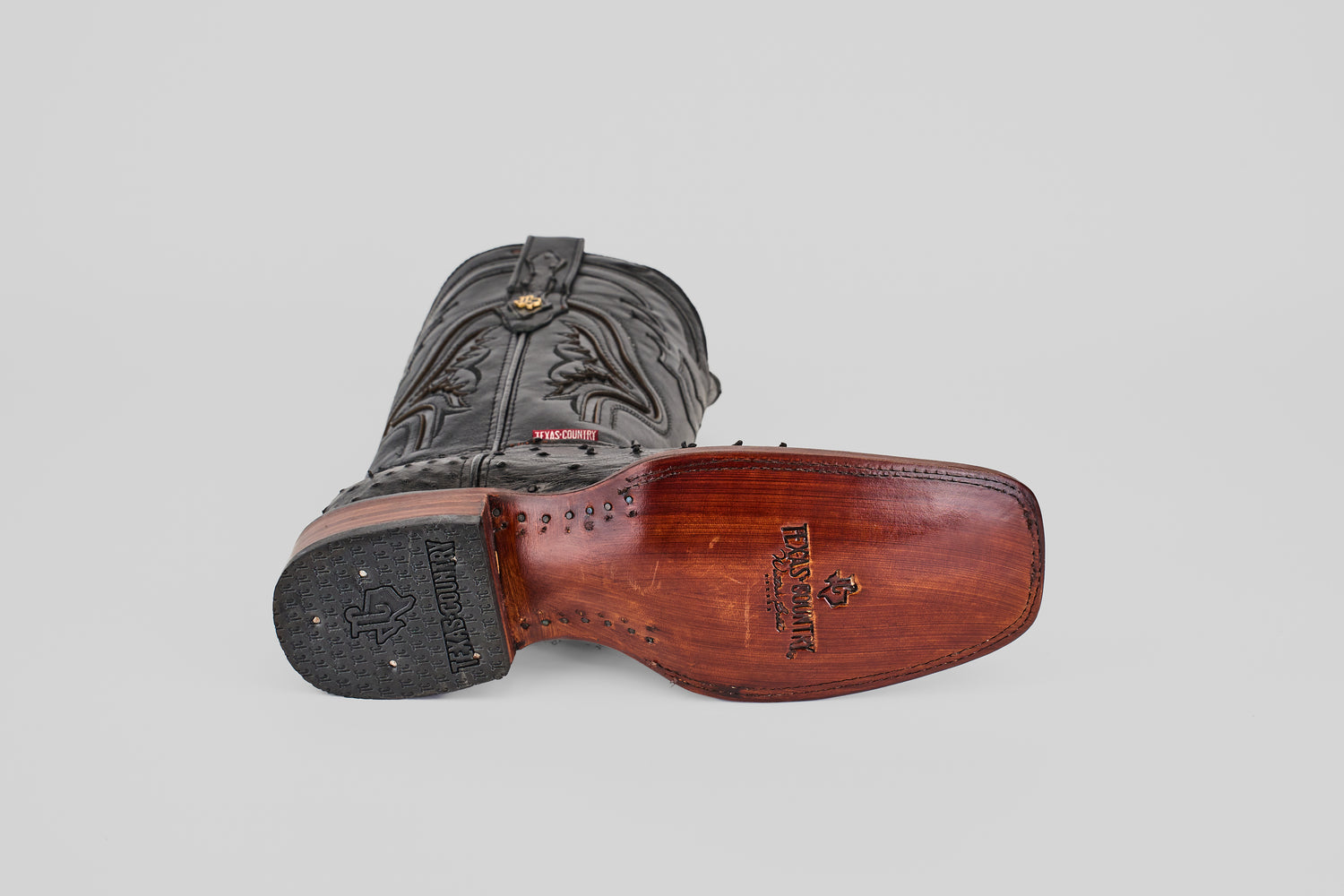 The Exotic Ostrich - Black - Rodeo Toe boot rests on its side, displaying its detailed brown leather sole with Tecovas branding and decorative stitching—an elegant piece of western wear.