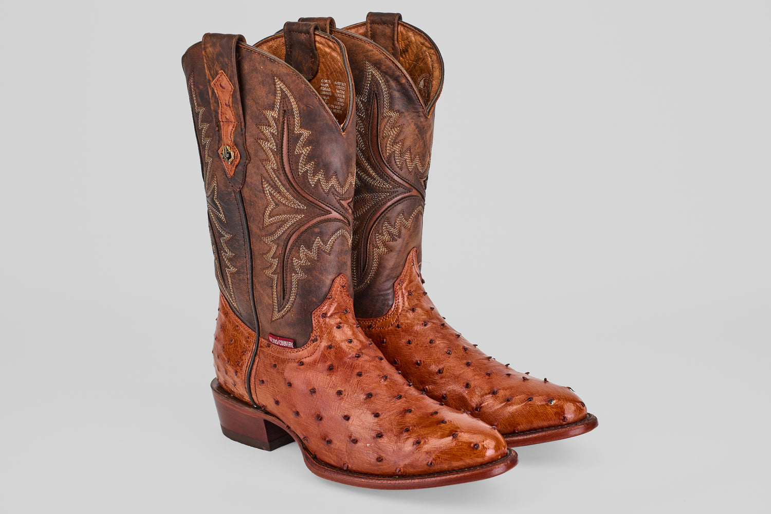 The Exotic Full Quilt Ostrich - Brandy - Round Toe boots feature handcrafted, textured ostrich leather with decorative stitching on tall, darker brown shafts, displayed against a plain light gray background.