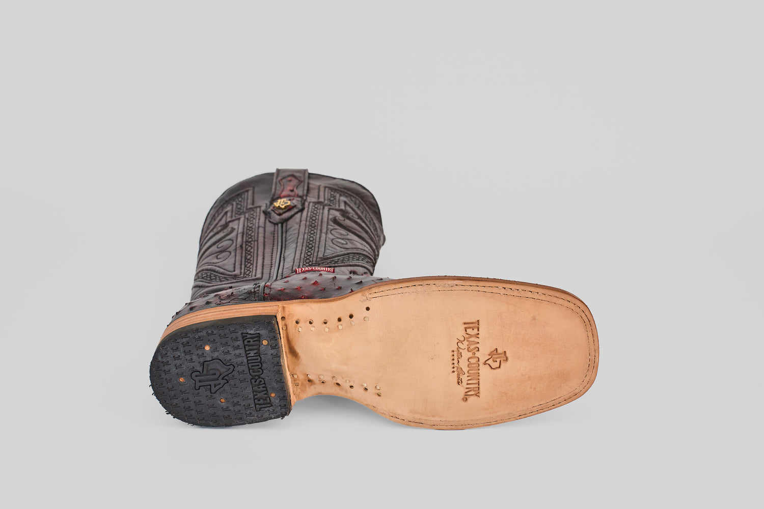 The Exotic Full Quilt Ostrich - Black Cherry - Square Toe cowboy boot rests on its side, featuring a leather sole and intricate upper stitching that exemplifies handcrafted artistry against a plain light background.