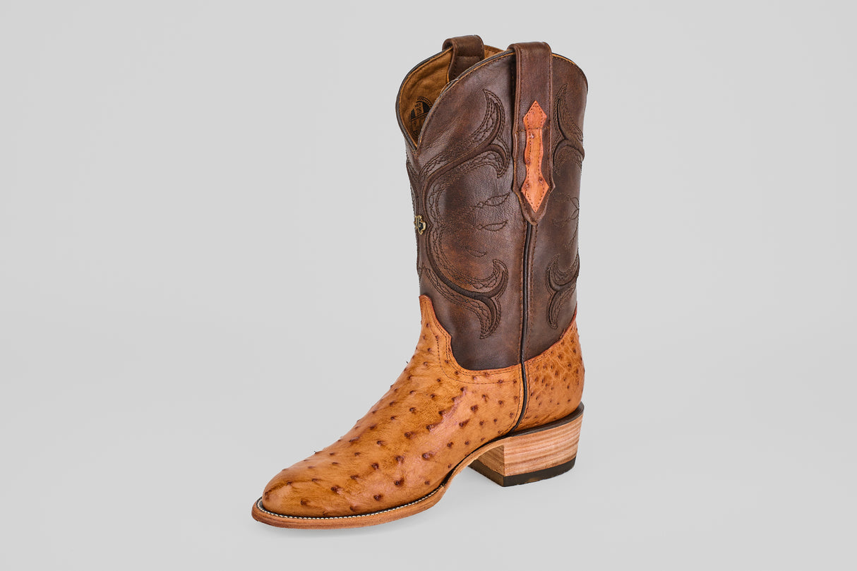 The Austin - Brandy - Roper Toe is a single men's cowboy boot with a tan ostrich leather foot, dark brown leather shaft, decorative stitching, and a stacked wooden heel, displayed on a plain gray background.