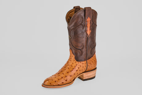 The Austin - Brandy - Roper Toe is a single men's cowboy boot with a tan ostrich leather foot, dark brown leather shaft, decorative stitching, and a stacked wooden heel, displayed on a plain gray background.