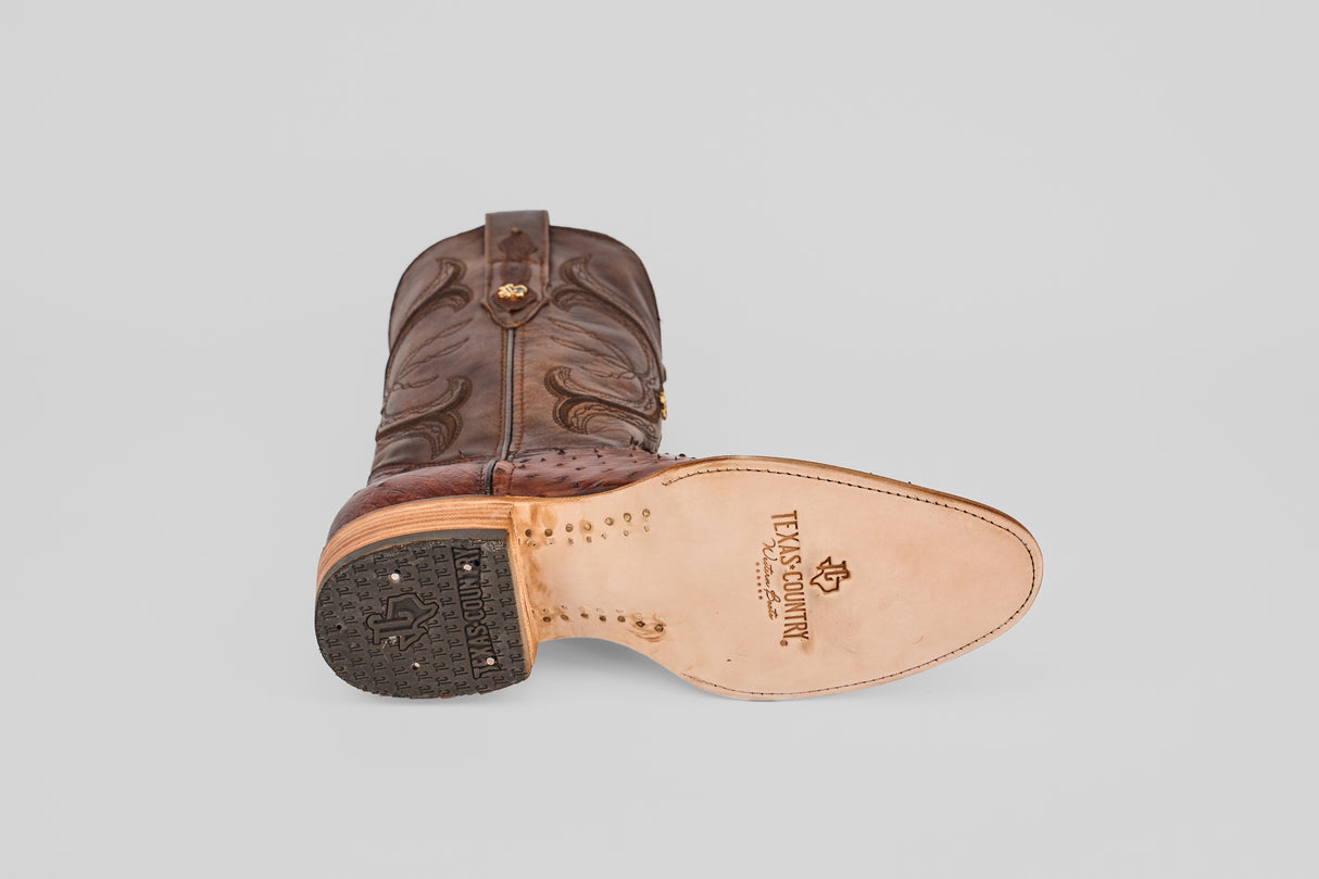 The Austin - Tabaco - Roper Toe men's cowboy boot, crafted from brown leather with decorative stitching, features a roper toe design, Texas Country logo, a sole on display, and a partially rubberized heel for extra traction.