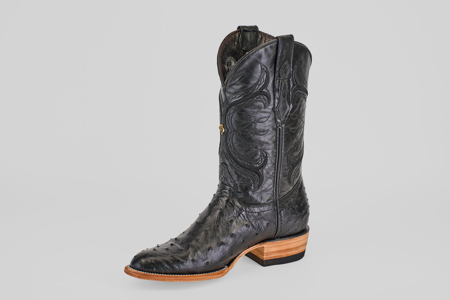 The Austin - Black - Roper Toe features black ostrich leather with intricate stitching, a textured surface, and a light brown stacked heel and sole—all set against a light gray background. Perfect for classic western boot lovers.