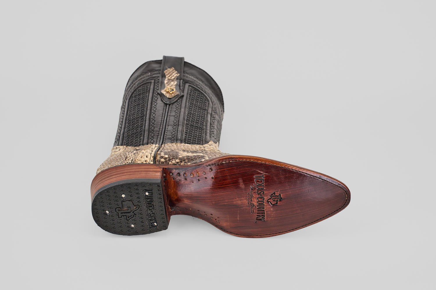 A single Tanner - Natural - Snip Toe cowboy boot rests on its side, featuring a textured black and tan Python Natural H-82 upper and wooden sole with leather finish, branding, and a rubber heel grip. The background is plain light gray.