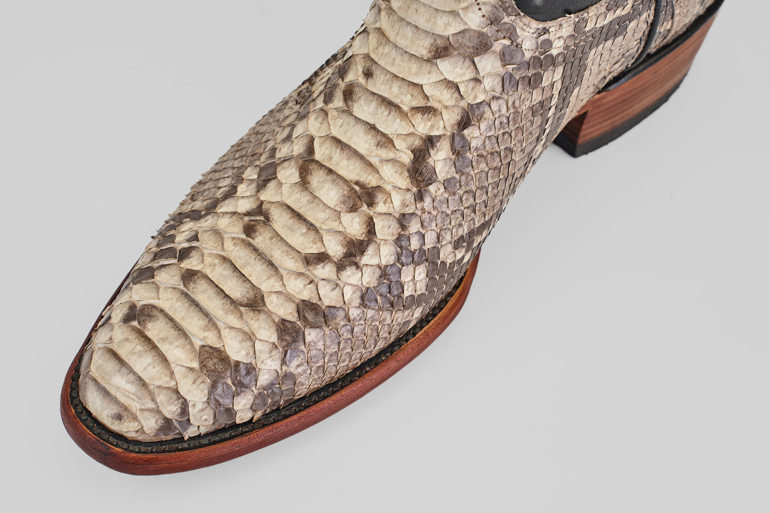 Close-up of the Tanner - Natural - Roper Toe boot’s pointed toe, crafted from natural beige and brown python leather, with a wooden sole and dark stitching detail.