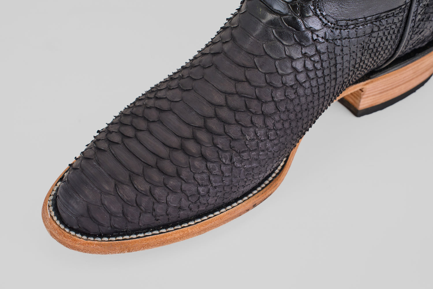 Close-up of the Tanner – Matte Black – Roper Toe boot, featuring a textured, scale-like leather pattern, wooden sole with light stitching, and photographed against a light gray background.