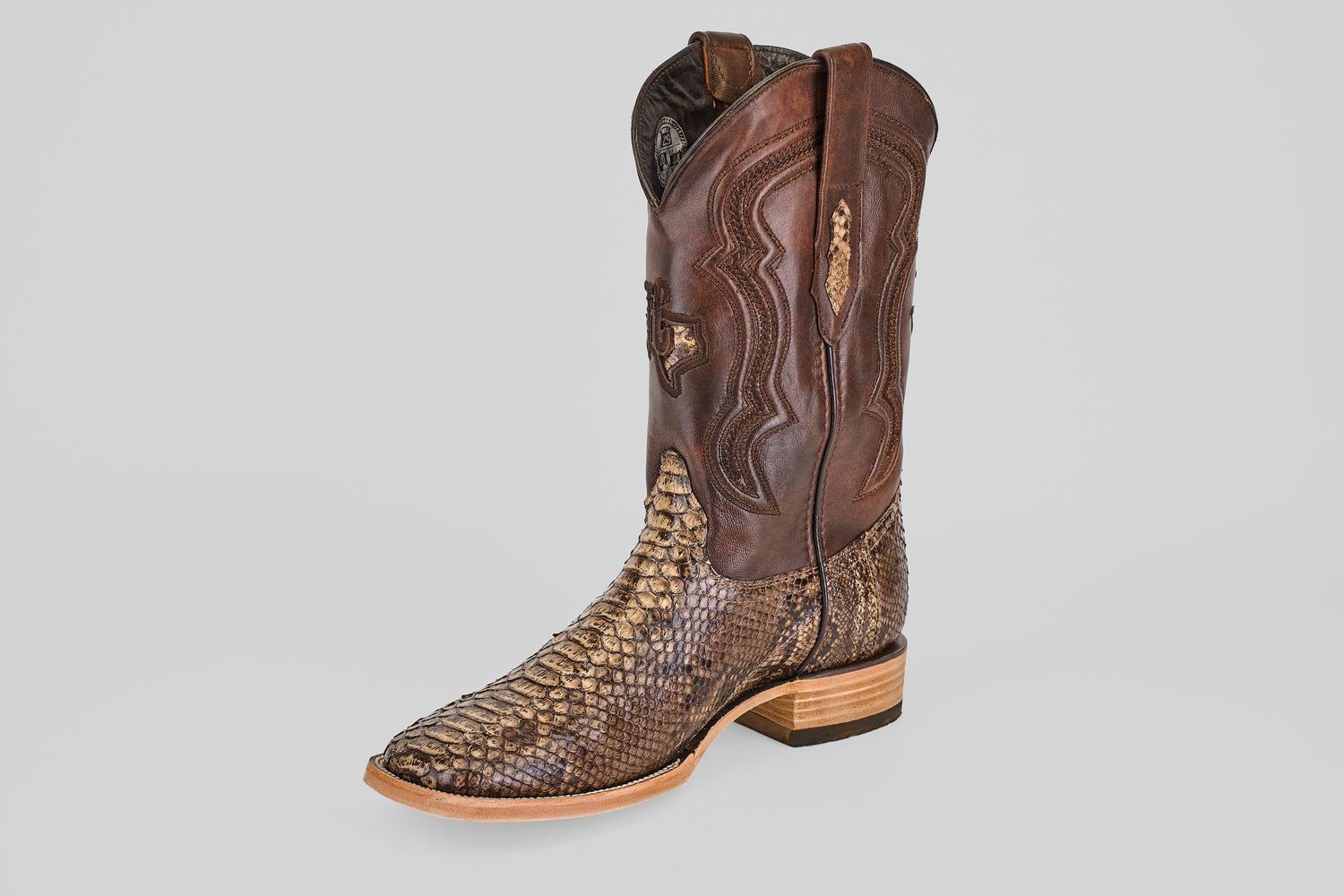The Tanner – Dune – Square Toe boot features a textured brown python leather lower half, a smooth darker brown leather upper with decorative stitching, and a wooden heel, displayed on a light gray background.
