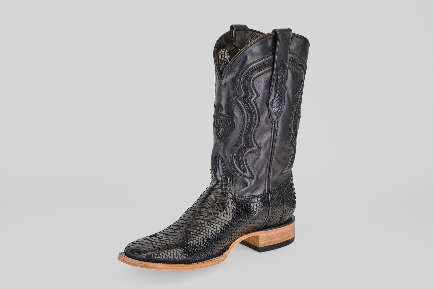 The Tanner - Black - Square Toe boot features a textured snakeskin pattern, intricate stitching on a tall shaft, and a light brown wooden heel and sole, shown against a plain light gray background.