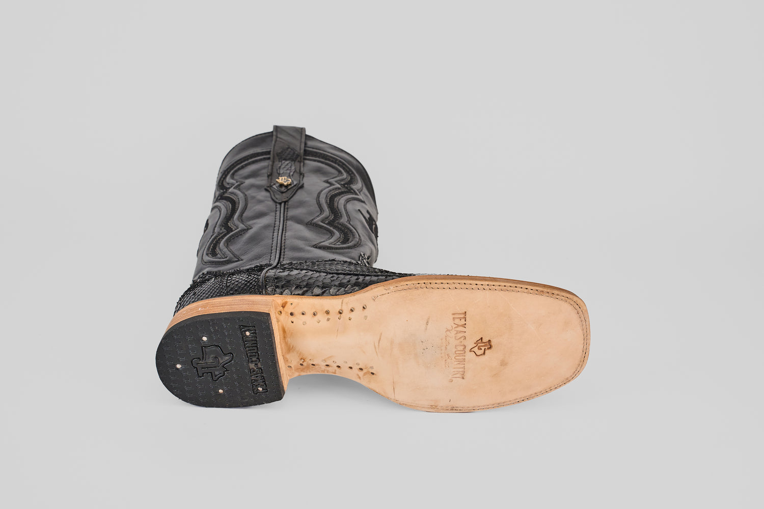 A single Tanner - Black - Square Toe boot lies on its side, showing the light tan sole with "TEXAS SCRIPT" and logo. The patterned black upper and treaded heel are clearly visible against a plain background.