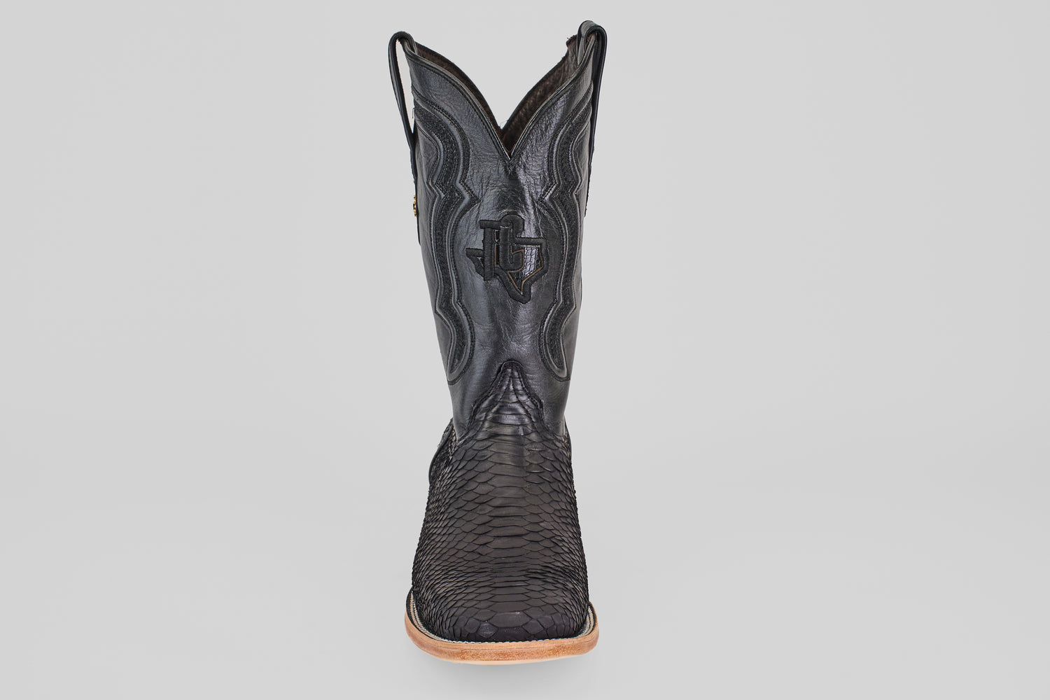 A single Tanner - Matte Black - Square Toe cowboy boot with a textured, snakeskin-inspired finish, matte black color, square toe, decorative stitching, and light brown sole is shown facing forward against a plain white background.