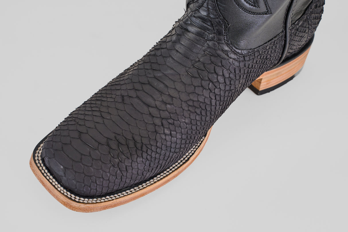 A close-up of the Tanner - Matte Black - Square Toe cowboy boot with python negro snakeskin and a light brown wooden sole, labeled PN30, set against a plain light background.