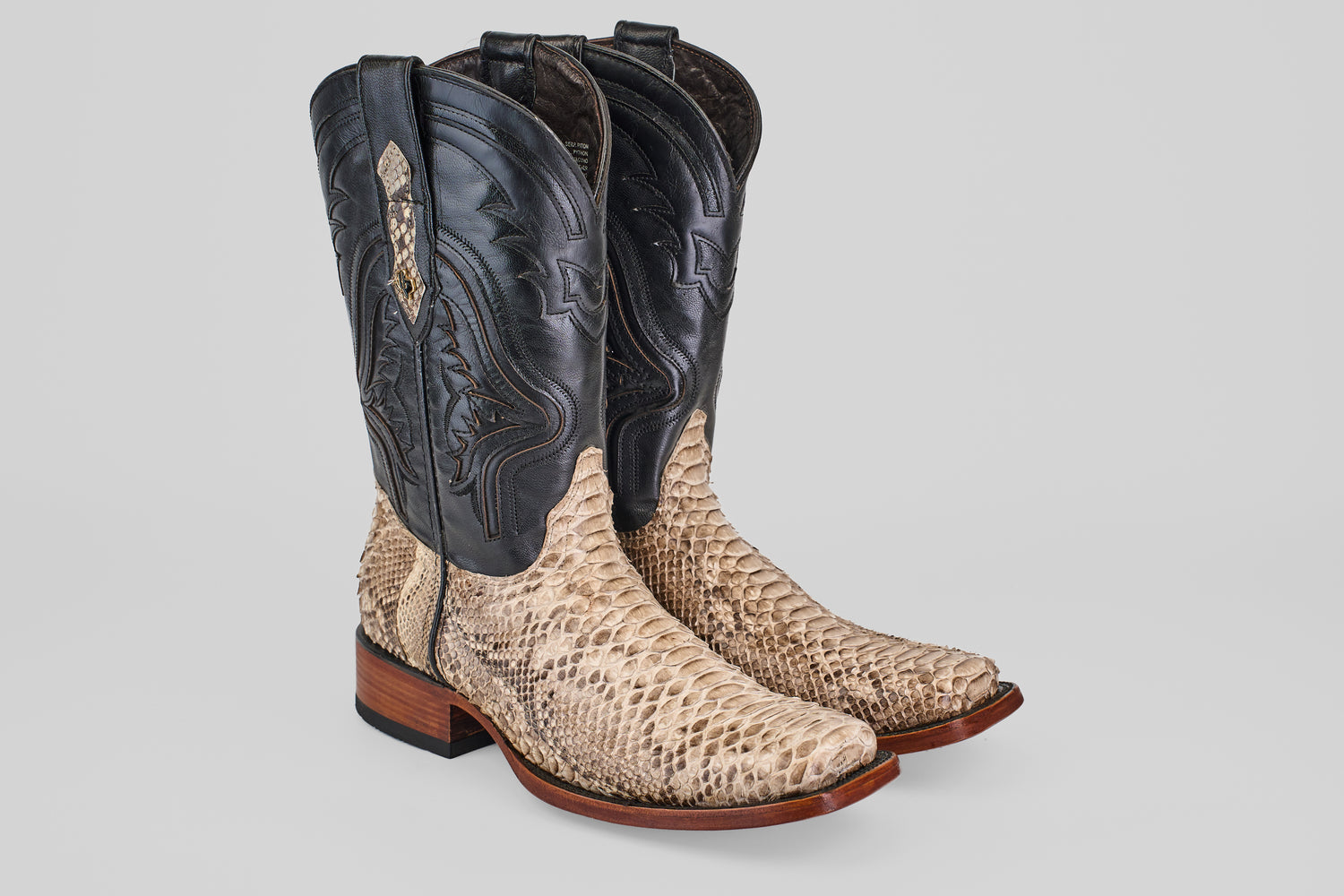 The Tanner - Natural - Rodeo Toe cowboy boots feature python natural leather on the foot, dark brown uppers with decorative stitching, and a wooden heel, shown on a light gray background—ideal for your next rodeo.
