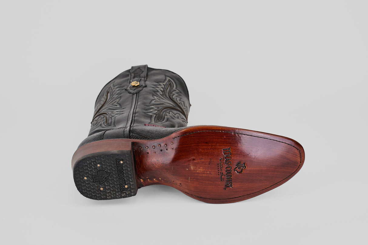 The Blaze – Black – Round Toe cowboy boot lies on its side, displaying a wooden sole with engraved brand logo and text, a nailed rubber heel, and intricate stitching on the leather upper. The round toe boot is set against a plain gray background.