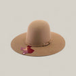 The 10x - Natural - TC Hats is a wide-brimmed tan felt cowboy hat with a decorative band and buckle, shown on a light background. A maroon Texas-shaped tag hangs from the brim.