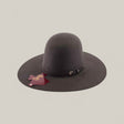 The 10X - Oxford - TC Hats is a wide-brimmed, dark brown Western hat with a shapeable crown and decorative strap, shown against a beige background. A maroon cattle brand-shaped tag hangs from the brim.