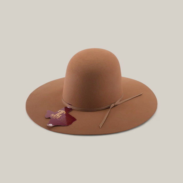 The 4X - Fawn - TC Hats is a light brown felt Western hat with a wide brim, round crown, and thin matching hatband. A maroon Africa-shaped tag hangs from the band. The hat is shown against a plain beige background.