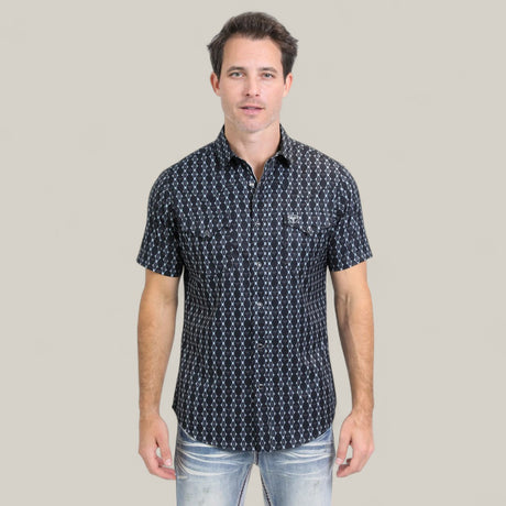 A man with short dark hair wears the Mens Performance Western Short Sleeve Aztec Print Navy Shirt (PGW10646) and light blue jeans, standing facing forward against a plain light beige background.