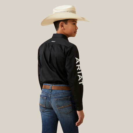 A young boy stands sideways in an Ariat Boys Black & White Team Logo Embroidered Long Sleeve Western Shirt (10045426), featuring a logo and white lettering on the sleeve and back. Hes paired it with blue jeans, a brown belt, and a wide-brimmed white cowboy hat, looking left against a plain white background.