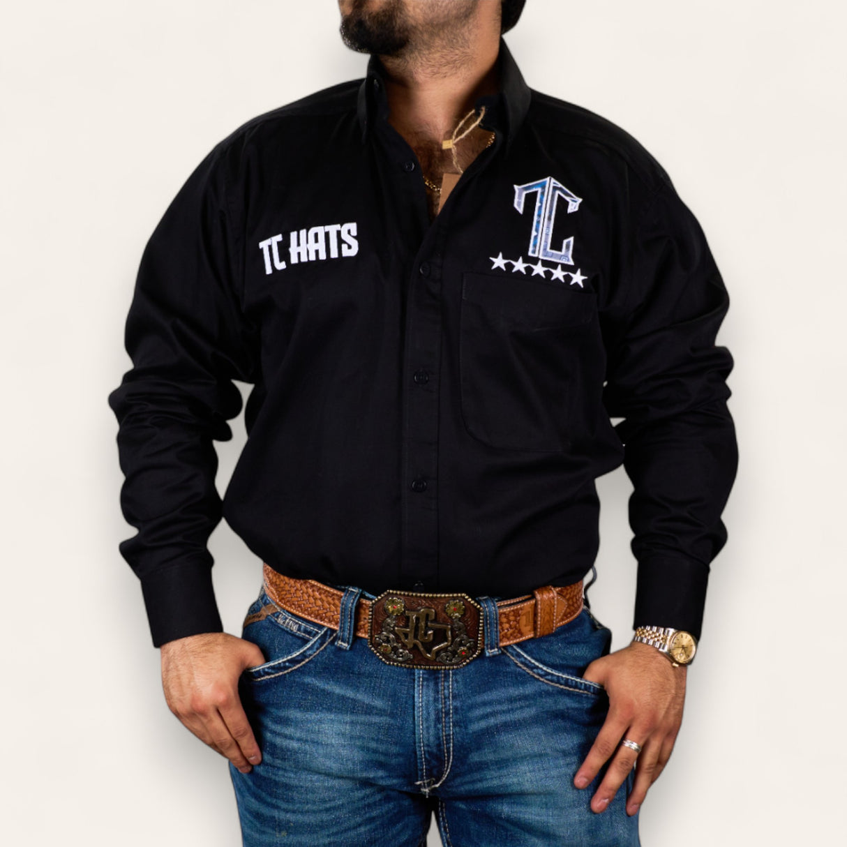 A man in a black button-up, blue jeans, brown woven belt with a large TC Aztec buckle, gold watch and necklace wears the TC Hats - Black Blue Aztec, featuring a white and blue logo and star, standing with hands in his pockets.